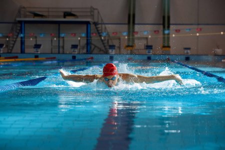 Téléchargez les photos : Nageur compétitif en bonnet rouge et lunettes en mouvement, technique de course de papillon d'entraînement, natation en piscine. Concept de sport professionnel, santé, endurance, force, mode de vie actif - en image libre de droit