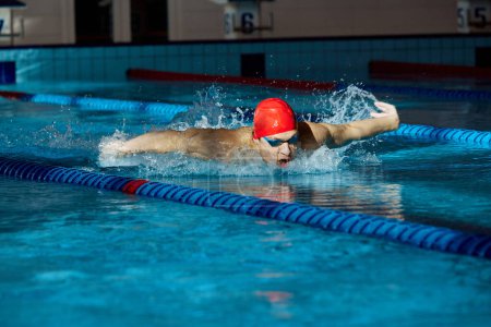 Foto de Joven con gorra roja y gafas, nadador en movimiento, mostrando un golpe de mariposa, entrenamiento, nadando en la piscina en el interior. Concepto de deporte profesional, salud, resistencia, fuerza, estilo de vida activo - Imagen libre de derechos