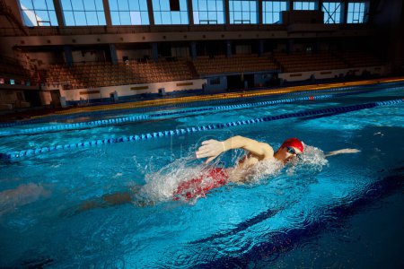 Téléchargez les photos : Jeune homme, nageur en bonnet rouge et lunettes en mouvement, entraînement coup libre, nager dans la piscine à l'intérieur. Concept de sport professionnel, santé, endurance, force, mode de vie actif - en image libre de droit