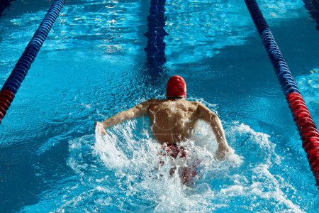 Téléchargez les photos : Vue de dessus du jeune homme musclé et athlétique, nageur en casquette rouge en mouvement, montrant la force, l'entraînement, la natation dans la piscine à l'intérieur. Concept de sport professionnel, santé, endurance, mode de vie actif - en image libre de droit