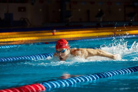 Foto de Joven musculoso mostrando espíritu competitivo, nadador en gorra y gafas de entrenamiento, nadando en piscina, preparándose para la competencia. Concepto de deporte profesional, salud, resistencia, estilo de vida activo - Imagen libre de derechos