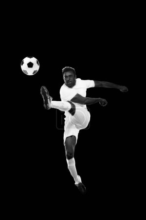 Téléchargez les photos : Image dynamique monochrome du jeune homme, footballeur en mouvement frappant le ballon dans un saut sur fond noir. Concept de sport professionnel, jeu, compétition, tournoi, action, mode de vie actif - en image libre de droit