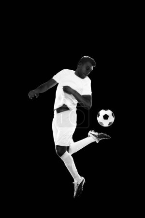 Photo for Concentrated young man, soccer player in white uniform training, dribbling ball isolated on black background. Concept of professional sport, game, competition, tournament, action, active lifestyle - Royalty Free Image
