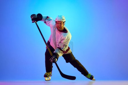 Foto de Hombre competitivo, motivado, jugador de hockey posando con palo, en uniforme y casco contra el fondo azul degradado en luz de neón. Concepto de deporte profesional, competición, juego, torneo - Imagen libre de derechos