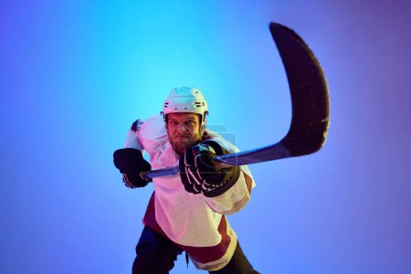 Téléchargez les photos : Homme compétitif et motivé, joueur de hockey posant avec bâton, en uniforme et casque sur fond bleu dégradé sous la lumière du néon. Concept de sport professionnel, compétition, jeu, tournoi - en image libre de droit