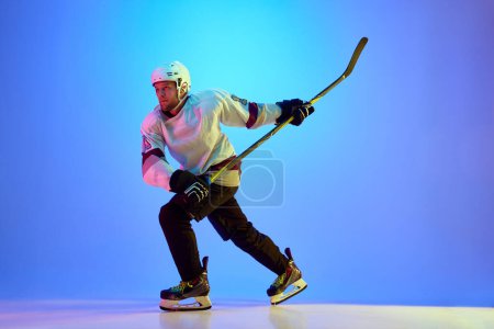 Téléchargez les photos : Homme, joueur de hockey portant uniforme et casque, posant avec un bâton sur fond bleu dégradé sous la lumière du néon. Concept de sport professionnel, compétition, jeu, tournoi - en image libre de droit