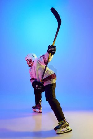 Téléchargez les photos : Homme ambitieux, joueur de hockey posant avec un bâton, montrant sa détermination contre un fond bleu dégradé sous la lumière du néon. Concept de sport professionnel, compétition, jeu, tournoi - en image libre de droit