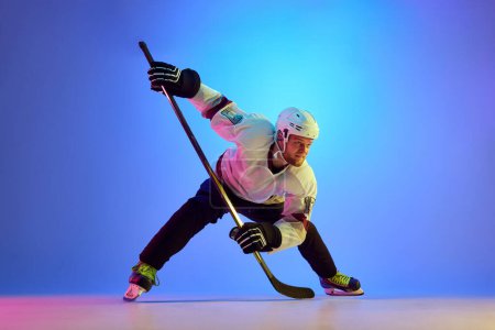 Téléchargez les photos : Homme ambitieux, joueur de hockey posant avec un bâton, montrant sa détermination contre un fond bleu dégradé sous la lumière du néon. Concept de sport professionnel, compétition, jeu, tournoi - en image libre de droit