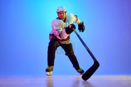 Téléchargez les photos : Homme compétitif et concentré, le joueur de hockey se dit prêt à gagner contre fond bleu dégradé sous la lumière du néon. Concept de sport professionnel, compétition, jeu, tournoi - en image libre de droit