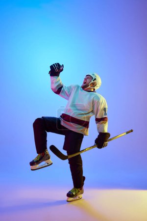 Foto de Hombre, jugador de hockey en casco, uniforme y palo expresando emociones de victoria contra el fondo azul degradado en luz de neón. Concepto de deporte profesional, competición, juego, torneo - Imagen libre de derechos
