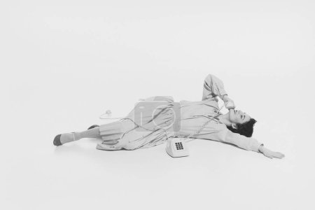 Young woman lying on floor and talking on retro style phone, smiling, laughing, Relaxed conversation. Monochrome image. Concept of retro and vintage, fashion, human emotions, lifestyle