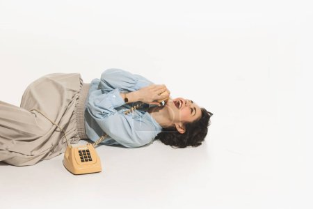 Happy, emotional, young woman lying on floor and talking on retro style phone, having fun on conversation, isolated on white. Concept of retro and vintage, fashion, human emotions, lifestyle