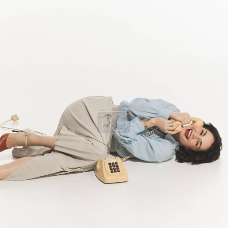Young beautiful woman with retro makeup, lying on floor and talking on vintage phone with happiness and joy, isolated on white. Concept of retro and vintage, fashion, human emotions, positive mood