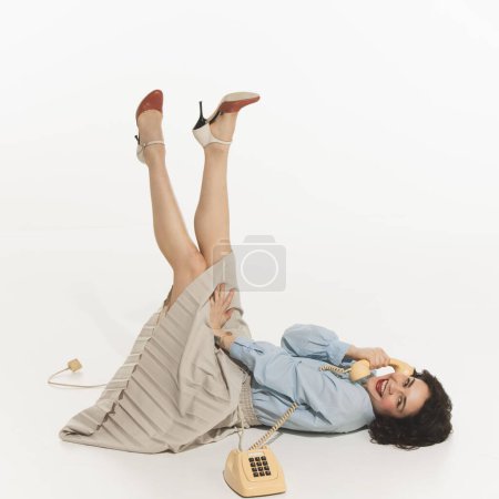 Mujer joven feliz, relajada y positiva tumbada en el suelo y hablando por teléfono de estilo retro, sonriendo, coqueteando, aislada sobre fondo blanco. Concepto de retro y vintage, moda, emociones humanas