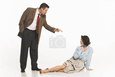 Téléchargez les photos : Homme en costume pointant vers une femme assise sur le sol, suggérant confrontation, dispute. Femme exprimant une réaction choquée aux affirmations de mans. Concept de rétro et vintage, mode, communication - en image libre de droit