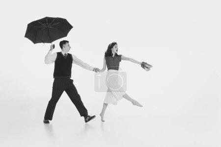 Photo for Charming young couple, dressed in vintage attire, holding hands and cheerfully walking under umbrella isolated on white background. Romantic date. Concept of retro and vintage, fashion, relationship - Royalty Free Image