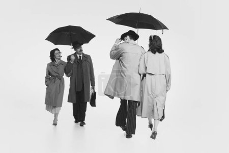 Téléchargez les photos : Deux couples en cloches élégantes, trench manteaux marchant sous le parapluie. Image monochrome. Salut en couple. Concept de rétro et vintage, mode, relation, amitié, communication - en image libre de droit