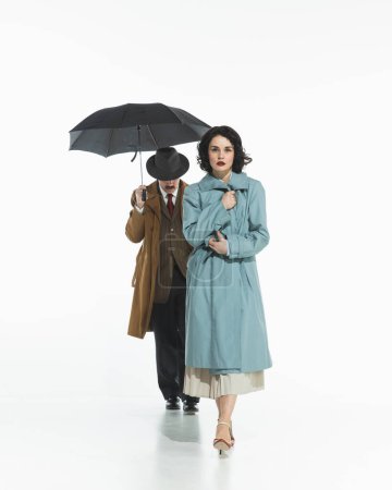 Téléchargez les photos : Élégante jeune femme en trench-coat bleu avec l'homme marchant derrière elle sous le parapluie. Sensations tendues, inquiétude intérieure. Concept de rétro et vintage, mode, relation, émotions humaines - en image libre de droit