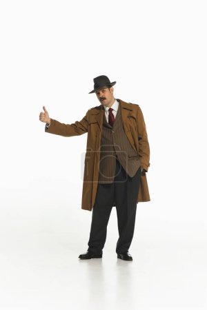 Man in a trench coat and fedora giving a thumbs up, suggesting hitchhiking, stopping car, taxi. Concept of retro and vintage, fashion, lifestyle, travelling
