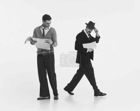 Téléchargez les photos : Deux hommes en tenue rétro avec des journaux isolés sur fond blanc. Image en noir et blanc. Un regard suspect. Concept de rétro et vintage, mode, émotions, affaires, style de vie - en image libre de droit