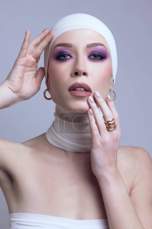 Téléchargez les photos : Portrait de belle jeune femme caucasienne maquillée, fard à paupières violet, touchant son visage en pose tendre, posant sur fond violet pastel. Concept de beauté, mode, cosmétique et cosmétologie - en image libre de droit