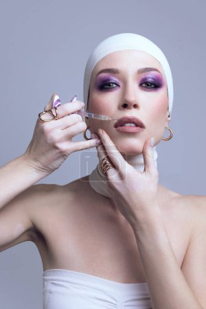 Young beautiful woman with bright makeup doing lip augmentation with syringe posing on pastel purple background. Concept of modern beauty standards, plastic surgery, health, cosmetology
