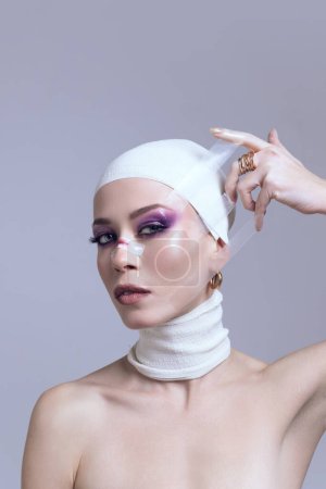 Frau mit hellem Augen-Make-up enthüllt Verband aus dem Gesicht nach einer plastischen Operation posiert auf pastellviolettem Hintergrund. Konzept moderner Schönheitsstandards, plastische Chirurgie, Gesundheit, Kosmetologie