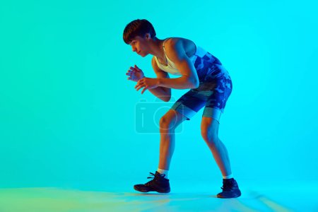 Athletic young man in blue uniform standing in wrestling pose, ready to fight against blue background in neon light. Concept of combat sport, martial arts, competition, tournament, athleticism