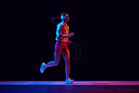 Photo for Young sportive woman keeping body in tone, running in comfortable sportswear and headphones on black background in neon light. Active and healthy lifestyle, sport, hobby, motivation, endurance concept - Royalty Free Image
