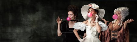 Elegant young women, medieval royal persons in period attire eating bubble gum against dark green background. Concept of comparison of eras, retro and vintage, history. Creative collage