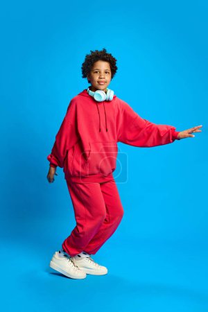 Foto de Chica afroamericana, niña con ropa casual escuchando música en auriculares, bailando y divirtiéndose contra el fondo azul del estudio. Concepto de infancia, emociones humanas, estilo de vida - Imagen libre de derechos