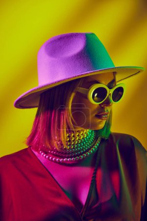 Photo for Avant-garde fashion portrait featuring model in hat and sunglasses against yellow background in neon light. Classic style. Concept of modern fashion, trendy style, beauty, youth culture - Royalty Free Image