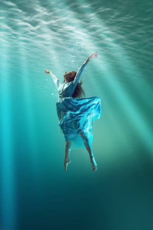 Photo for Under the sea surface. Young girl in elegant dress levitating underwater, extending hands toward sunlight filtering through water. Concept of surrealism, beauty, mystery and fantasy, freedom - Royalty Free Image