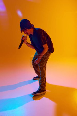 Photo for Dynamic performance. Young musician delivers dynamic rap, captivating the audience with creative art against yellow background in neon light. Concept of music, performance, festival, live concert - Royalty Free Image