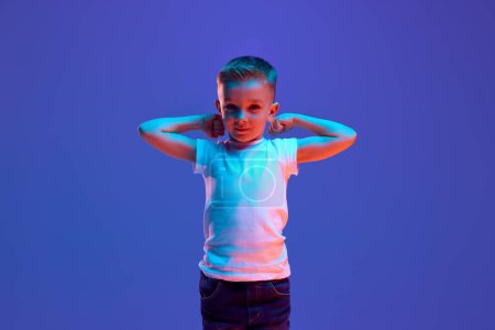 Photo for Portrait of little child, boy raising hands showing his power and playfully looking at camera in mixed neon light against blue background. Concept of fashion and style, beauty, back to school, - Royalty Free Image