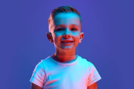 Photo for Portrait of charming child, boy dressed white T-shirt looking at camera widely smiling in mixed neon light against blue background. Concept of human emotions, fashion and style, beauty, back to school - Royalty Free Image