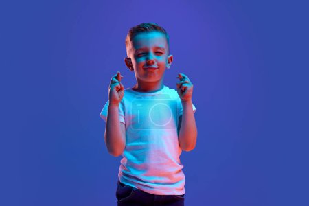 Photo for Good luck at exam. Cute child, boy crossed fingers and closed eyes to get lucky in mixed neon light against blue background. Concept of fashion and style, beauty, back to school, education, fortune. - Royalty Free Image