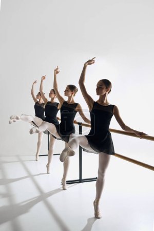 Foto de Cuatro chicas, bailarinas de ballet en el estudio de pie en la barra, extendiéndose con gracia con un brazo, practicando sobre fondo gris. Concepto de ballet, arte, estudio de danza, estilo clásico, juventud - Imagen libre de derechos