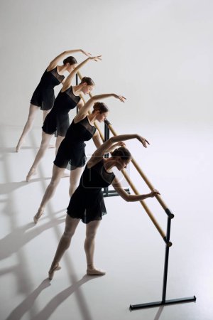 Foto de Simetría y gracia en el ballet. Cuatro chicas flexibles, bailarinas de ballet de pie en la barra y entrenamiento contra el fondo gris del estudio. Concepto de ballet, arte, estudio de danza, estilo clásico, juventud - Imagen libre de derechos