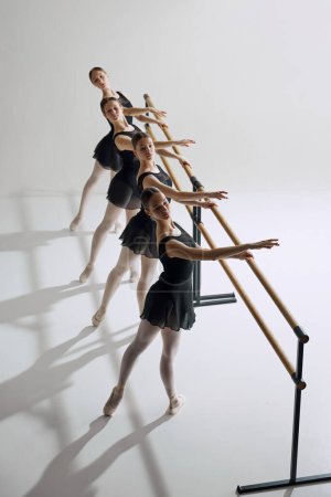 Téléchargez les photos : Quatre adolescentes, danseuses de ballet pratiquant à la barre, étendant le bras en geste délicat sur fond de studio gris. Concept de ballet, art, studio de danse, style classique, jeunesse - en image libre de droit