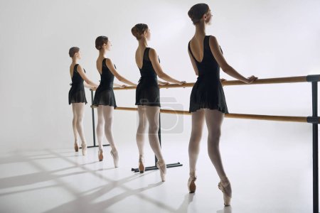 Photo for Four ballet dancers, elegant teen girls standing at barre in black costumes and pointe shoes, practicing against grey studio background. Concept of ballet, art, dance studio, classical style, youth - Royalty Free Image