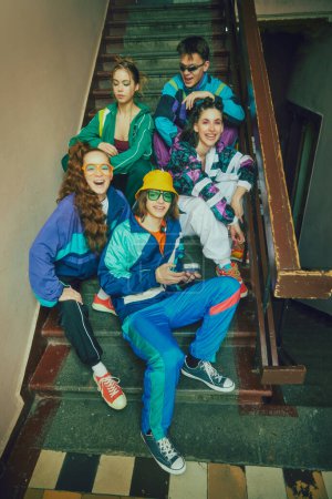 Photo for Vibrant youth atmosphere. Young boys and girls, friends wearing colorful 90s style sportswear, posing on stairs, entryway. Concept of 90s, fashion, youth culture, old-style trends - Royalty Free Image