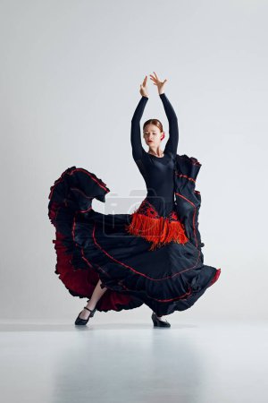 Photo for Artistic woman, flamenco dancer n striking black and red dress performing with passion, dancing against grey studio background. Concept of art of movement, classical dance, beauty, festival - Royalty Free Image