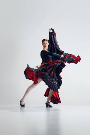 Téléchargez les photos : Capture artistique de la danseuse de flamenco en mouvement, élégante femme en robe noire et rouge faisant des mouvements dynamiques sur fond de studio gris. Concept d'art du mouvement, danse classique, beauté, festival - en image libre de droit