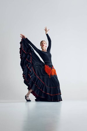 Photo for Grace of flamenco dance. Artistic talented woman in stylish black and red dress performing flamenco against grey studio background. Concept of art of movement, classical dance, beauty, festival - Royalty Free Image
