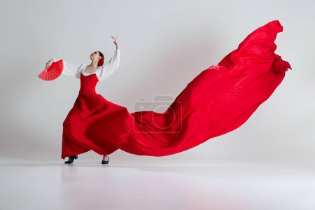 Téléchargez les photos : Femme danseuse de flamenco en mouvement, femme élégante avec costume rouge et éventails exécutant la danse flamenco sur fond de studio gris. Concept d'art du mouvement, danse classique, beauté, festival - en image libre de droit