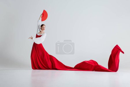 Foto de Bailarina flamenca femenina en movimiento, mujer elegante con traje rojo y abanicos bailando flamenco sobre fondo gris estudio. Concepto de arte del movimiento, danza clásica, belleza, festival - Imagen libre de derechos