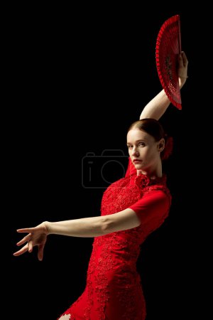 Photo for Flamenco dancer in elegant red dress and fan strikes captivating pose, highlighting rhythmic essence of traditional Spanish dance. Concept of art of movement, classical dance, beauty, festival - Royalty Free Image