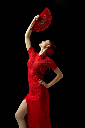 Foto de Bailarina flamenca en vestido rojo adornado de pie con elegancia, posando con abanico rojo sobre fondo negro. Concepto de arte del movimiento, danza clásica, belleza, festival - Imagen libre de derechos