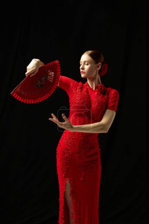 Photo for Flamenco female dancer in ornate red dress standing with elegance, posing with red fan against black background. Concept of art of movement, classical dance, beauty, festival - Royalty Free Image